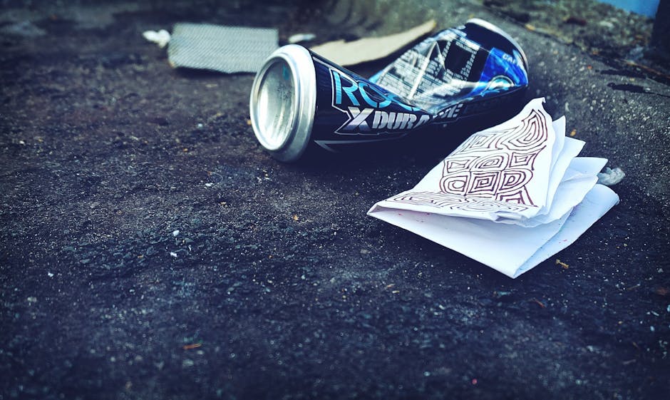 A crushed blue and black energy drink can with silver edges lies on a rough, dark asphalt surface, partially rolled onto its side. Next to the can, there is a crumpled piece of white paper with red ma