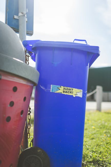 A crushed blue and black energy drink can with silver edges lies on a rough, dark asphalt surface, partially rolled onto its side. Next to the can, there is a crumpled piece of white paper with red ma