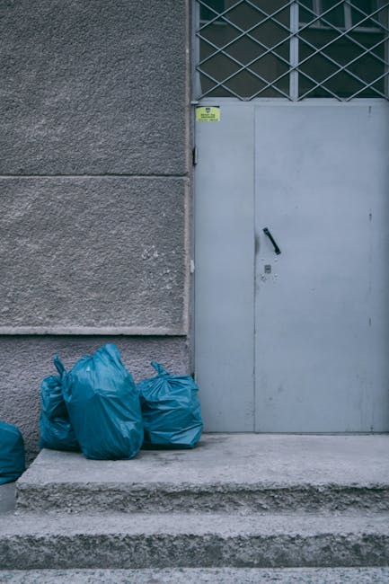 A large collection of overflowing rubbish bins and scattered waste bags placed on a paved sidewalk in front of a commercial building, with various types of waste including crumpled cardboard boxes, pl