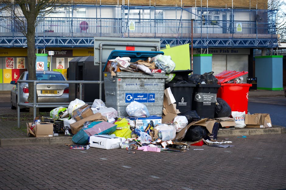In the foreground of the image, there is a large pile of mixed household rubbish overflowing from several black and grey wheelie bins labeled for paper, mixed waste, and general waste, with some lids